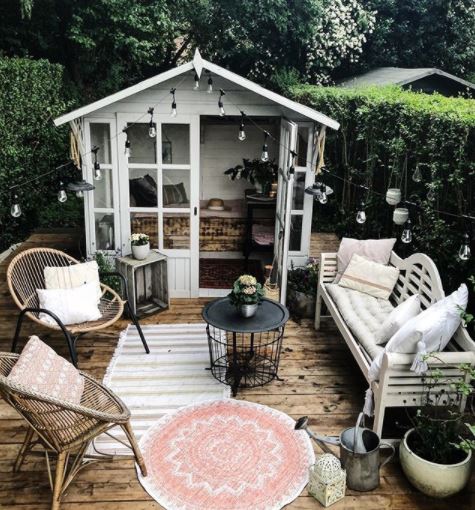 a pretty she shed with glass doors opens onto a deck with chairs, bench and outdoor rugsx