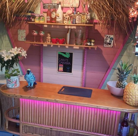 a tiki style shed bar with neon pink lights, kitschy decor and shelves filled with cocktail spirits