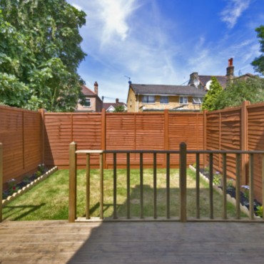 a simple rectangular garden with a deck and lawn with narrow floral borders and a sturdy looking fence