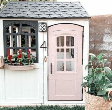 a grown-up playhouse for kids with a pastel pink door, picket fence and window box with flowers